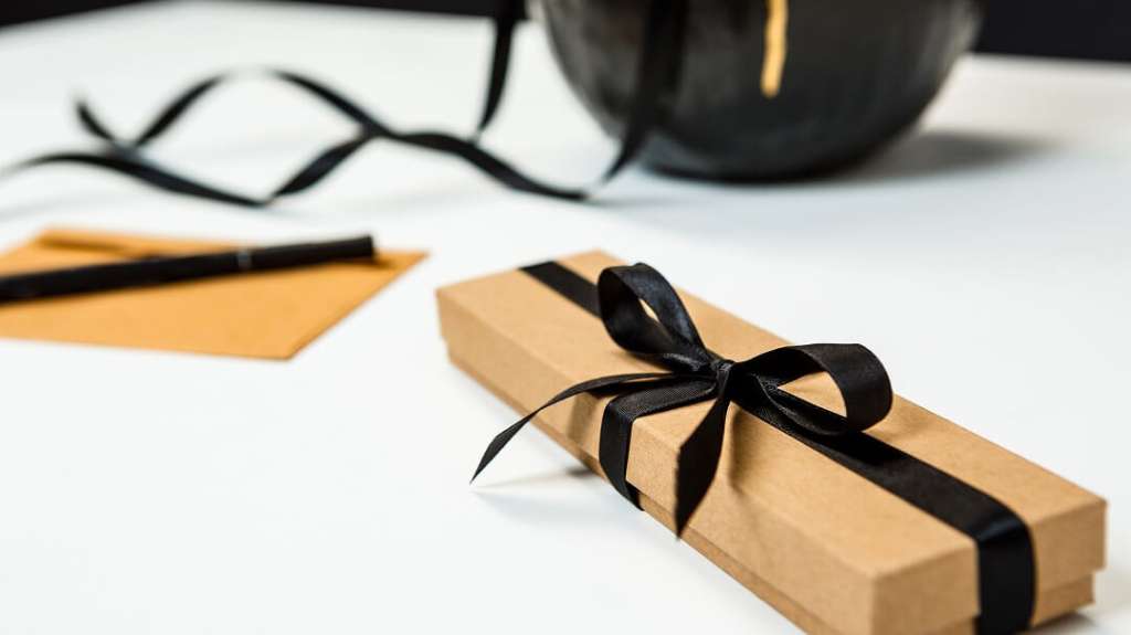 Un élégant coffret cadeau noir ouvert sur une surface sombre et texturée, présentant des cadeaux d'affaires haut de gamme, notamment un thermos épuré, un carnet, un stylo de qualité et des éléments botaniques secs décoratifs.