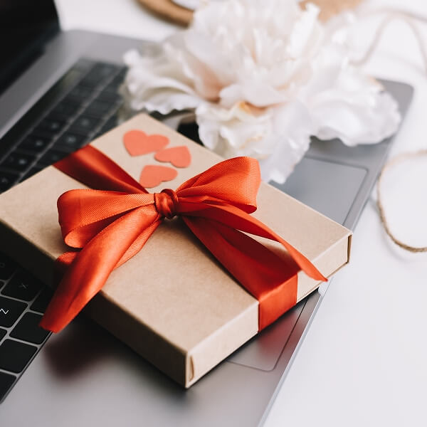 Un gros plan des mains d'une personne offrant une boîte cadeau en carton kraft joliment emballée avec un ruban blanc propre à une autre personne dans un cadre professionnel ou de bureau.